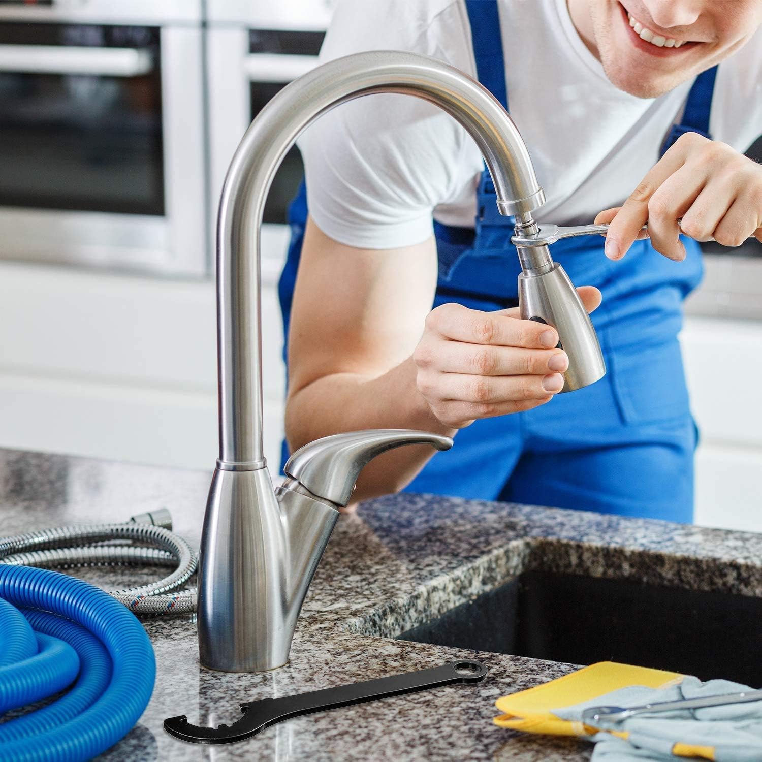 Man in blue overalls smiling while installing a stainless steel kitchen faucet. Tools and hoses are on the granite countertop, conveying a sense of repair work.