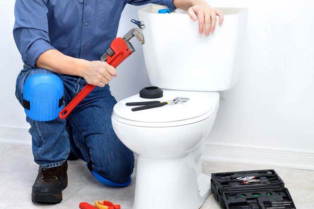 A person kneels by a toilet in a bathroom, holding a red wrench. Nearby, tools are arranged on the toilet lid and an open toolbox is on the floor.